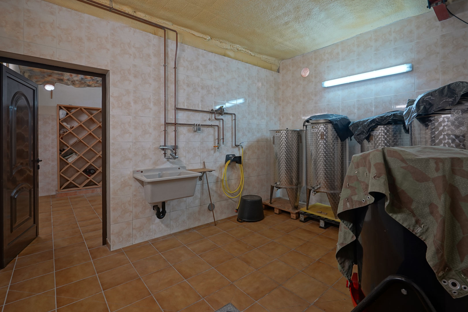 Utility room for wine production featuring stainless steel tanks, a deep utility sink with copper piping, and tiled walls. A wooden wine rack is visible in the adjacent room.