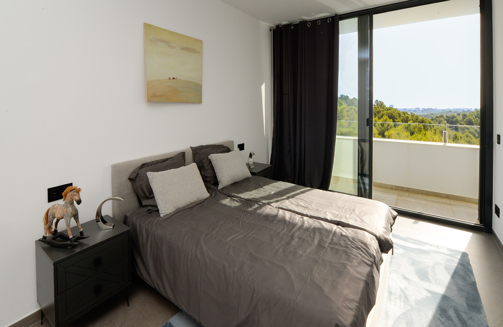 Bright bedroom featuring a double bed with charcoal linens, minimalist nightstands, and floor-to-ceiling glass doors opening onto a private terrace with nature views.