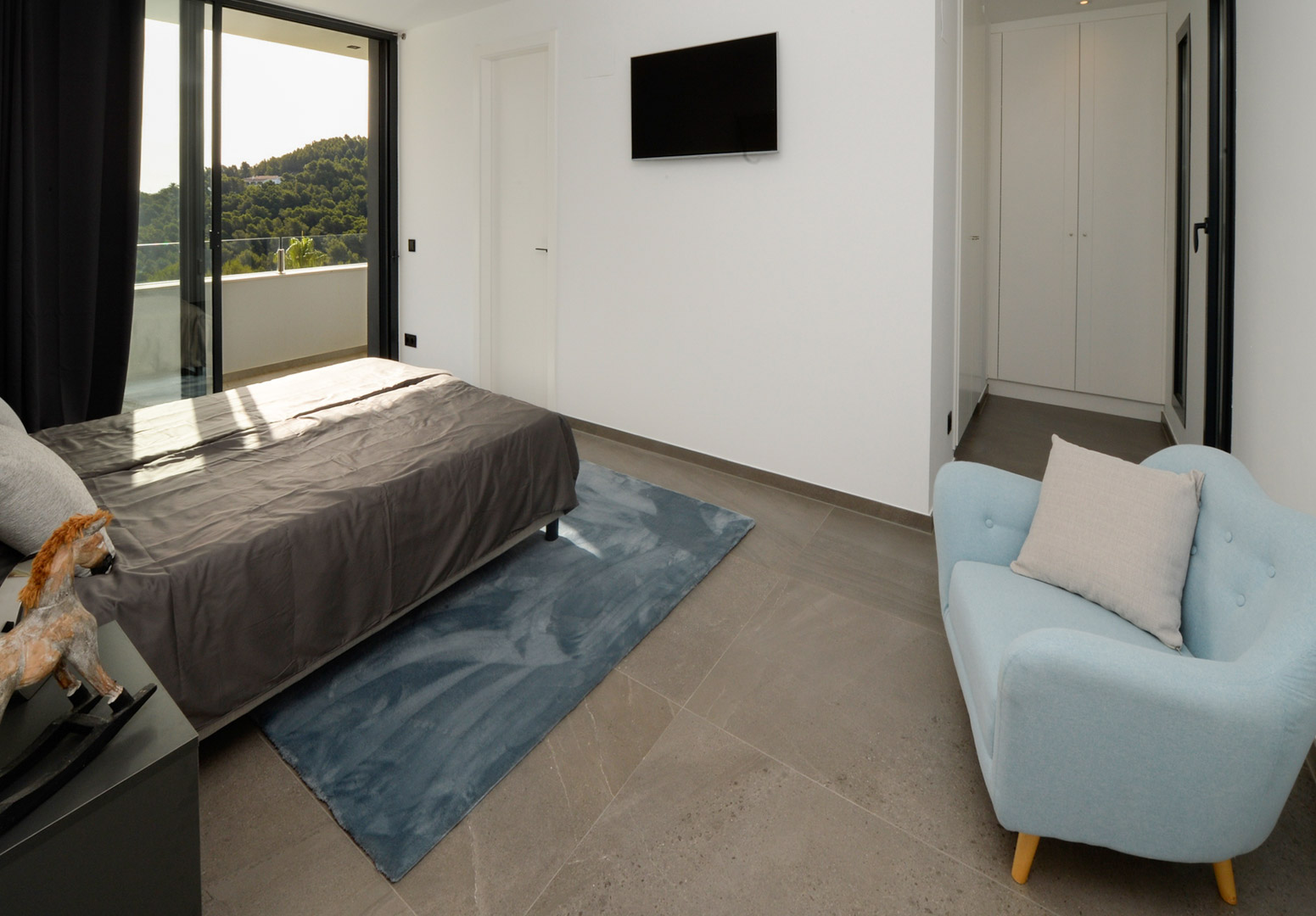 Contemporary bedroom with terrace access, grey stone-effect flooring, integrated white wardrobes, and panoramic views of the Mediterranean landscape.