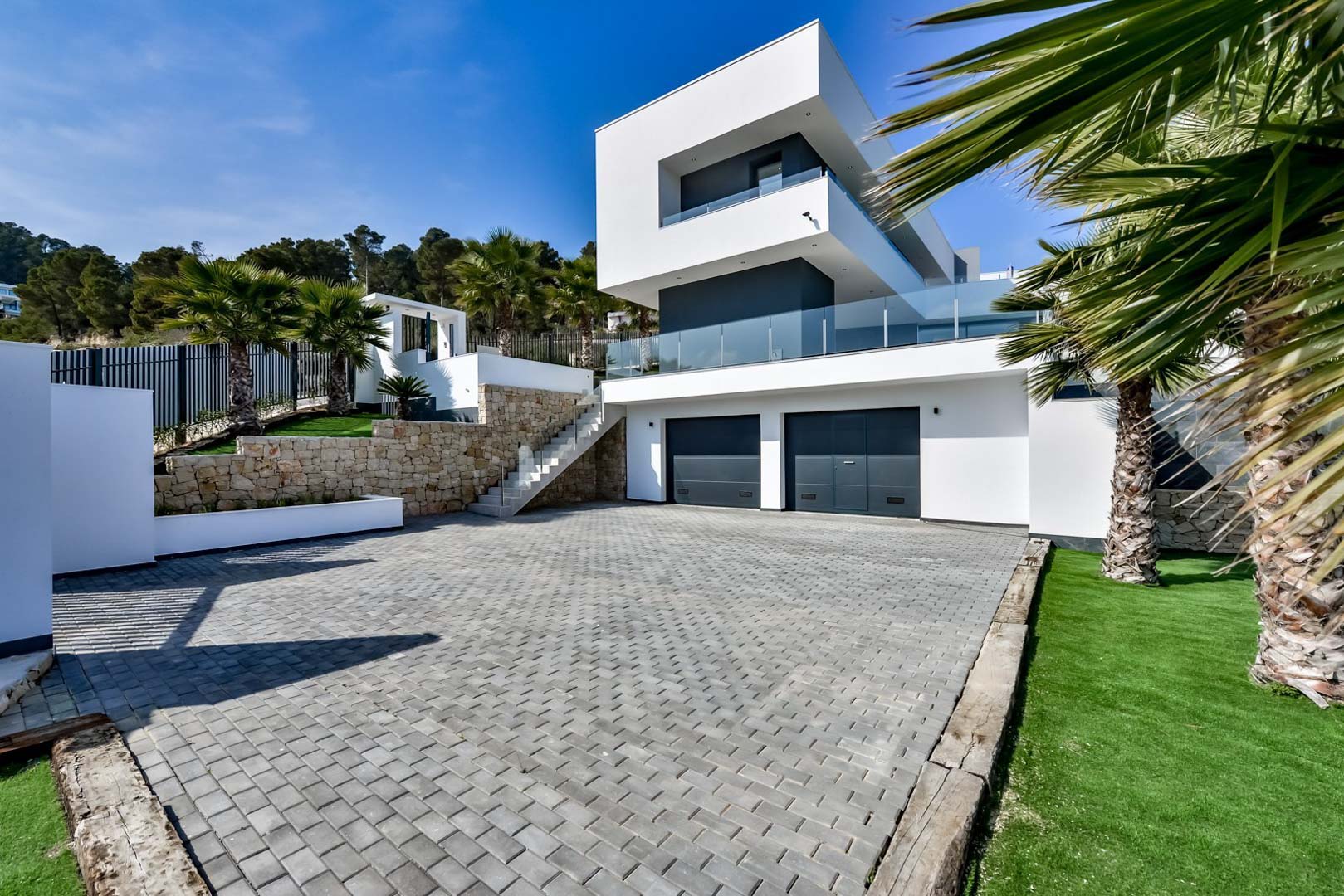 Contemporary exterior featuring a double garage, natural stone retaining walls, and a paved driveway framed by Mediterranean landscaping.