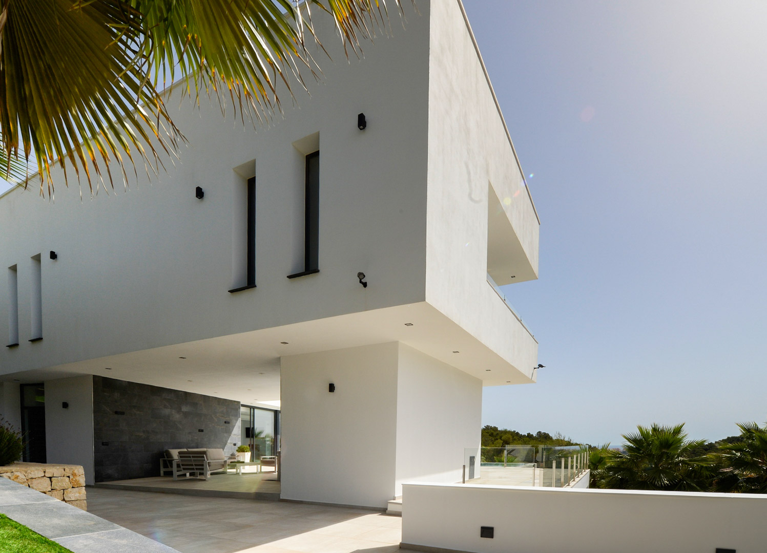 Contemporary facade featuring an architectural cantilever, grey natural stone walls, and a covered terrace with outdoor lounge furniture and recessed lighting.