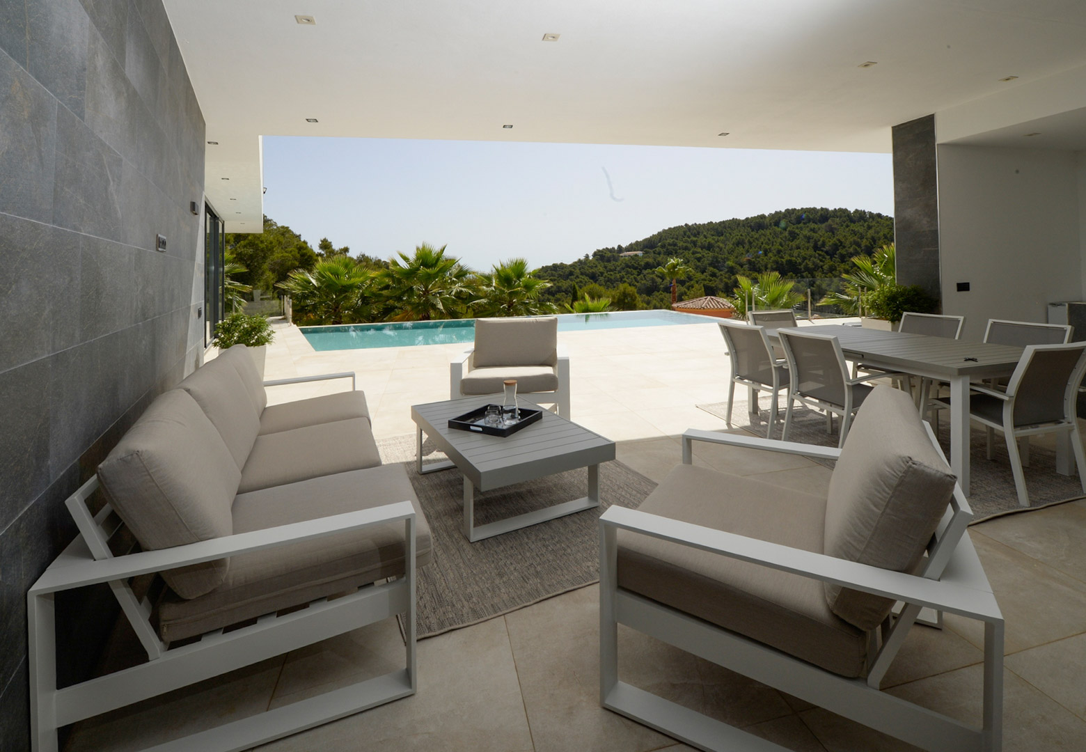 Covered terrace featuring a modern lounge and outdoor dining area, overlooking the infinity pool and lush Mediterranean hills.