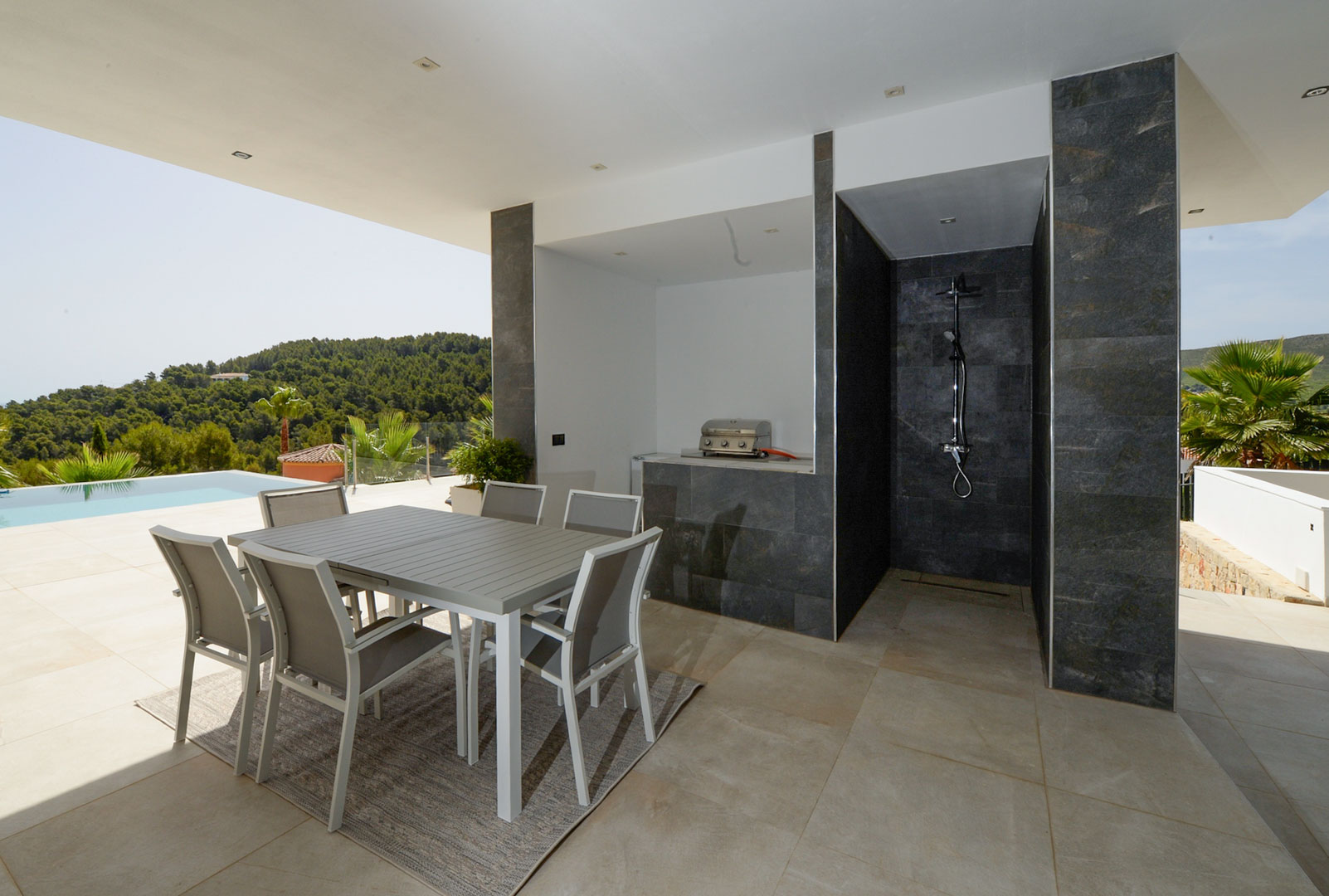 Covered terrace featuring an outdoor dining area, summer kitchen with barbecue, and a dark stone-clad outdoor shower overlooking the pool.
