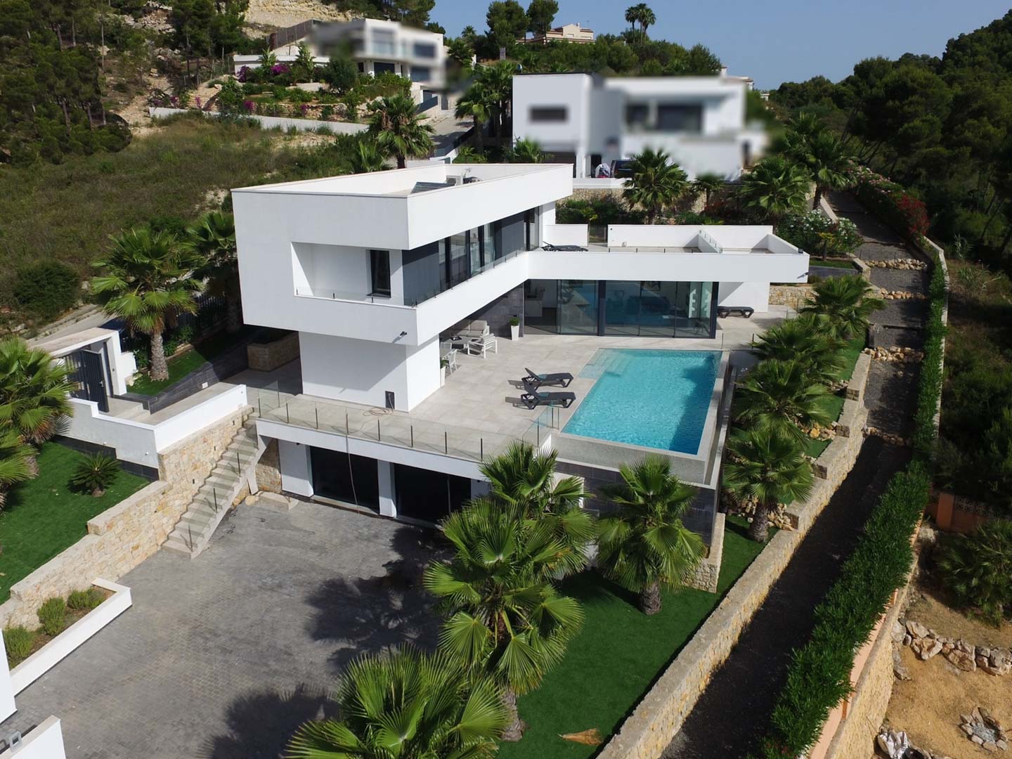 Elevated view of the modern villa featuring white geometric architecture, an infinity pool, stone-paved terraces, and a landscaped garden with palm trees.