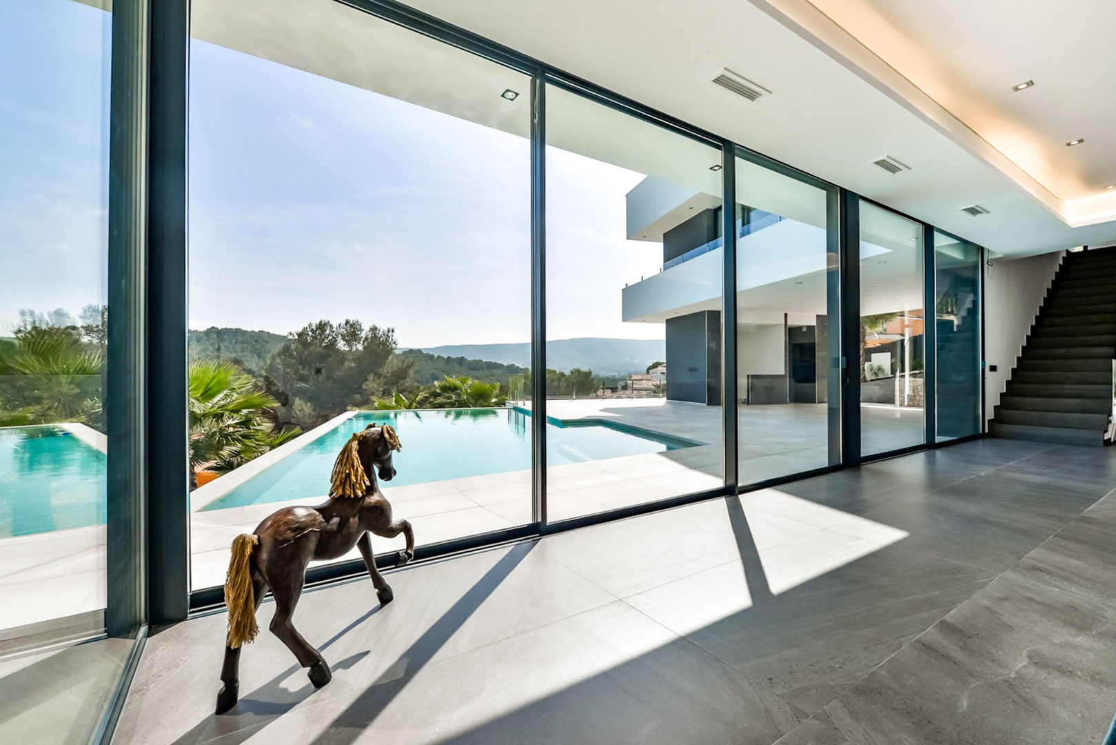 Living area featuring floor-to-ceiling glass walls that open to the infinity pool and terrace. Grey porcelain flooring and a modern staircase lead to the upper level.
