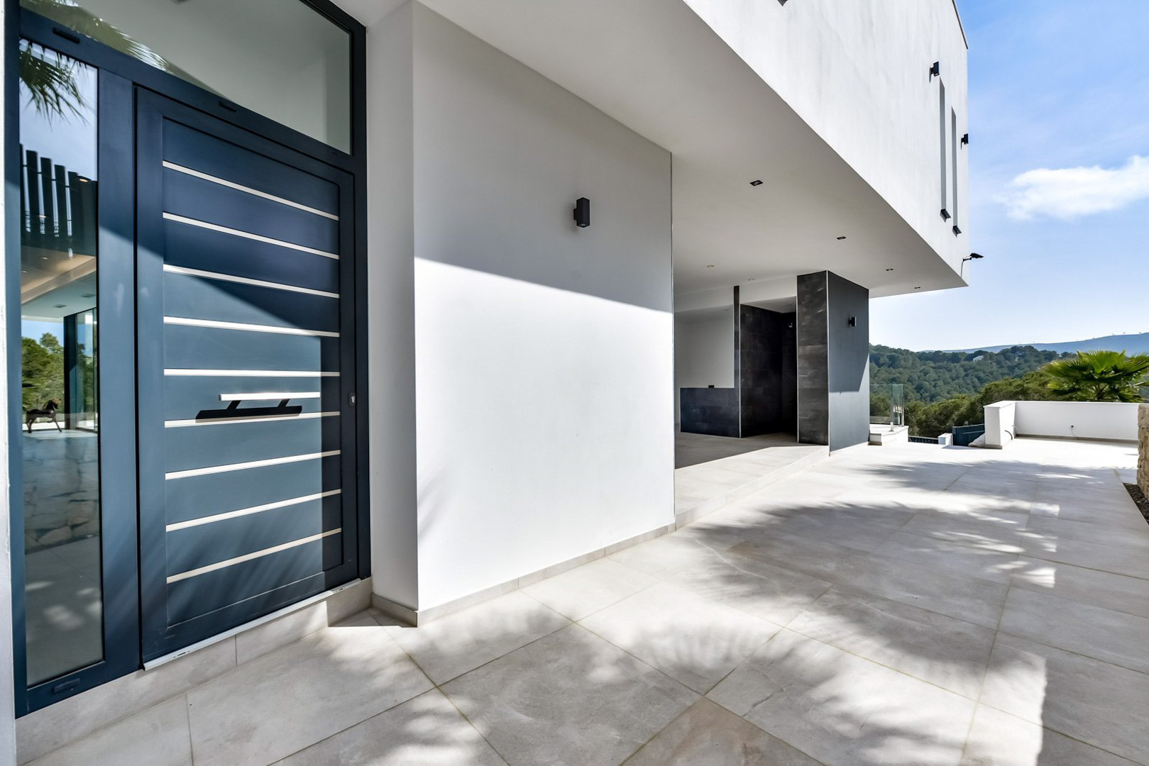 Main entrance featuring a modern dark blue door with metallic accents, a spacious tiled porch, and views of the surrounding green hills and Mediterranean landscape.