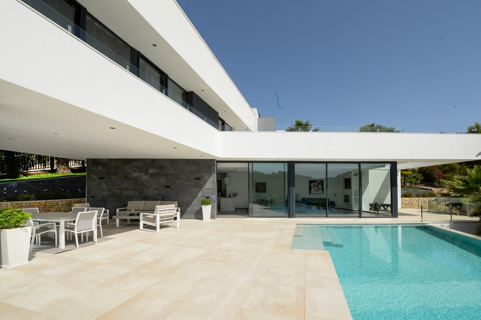 Main terrace featuring an infinity pool, covered outdoor dining area, and minimalist white architecture with floor-to-ceiling glass doors and stone accents.