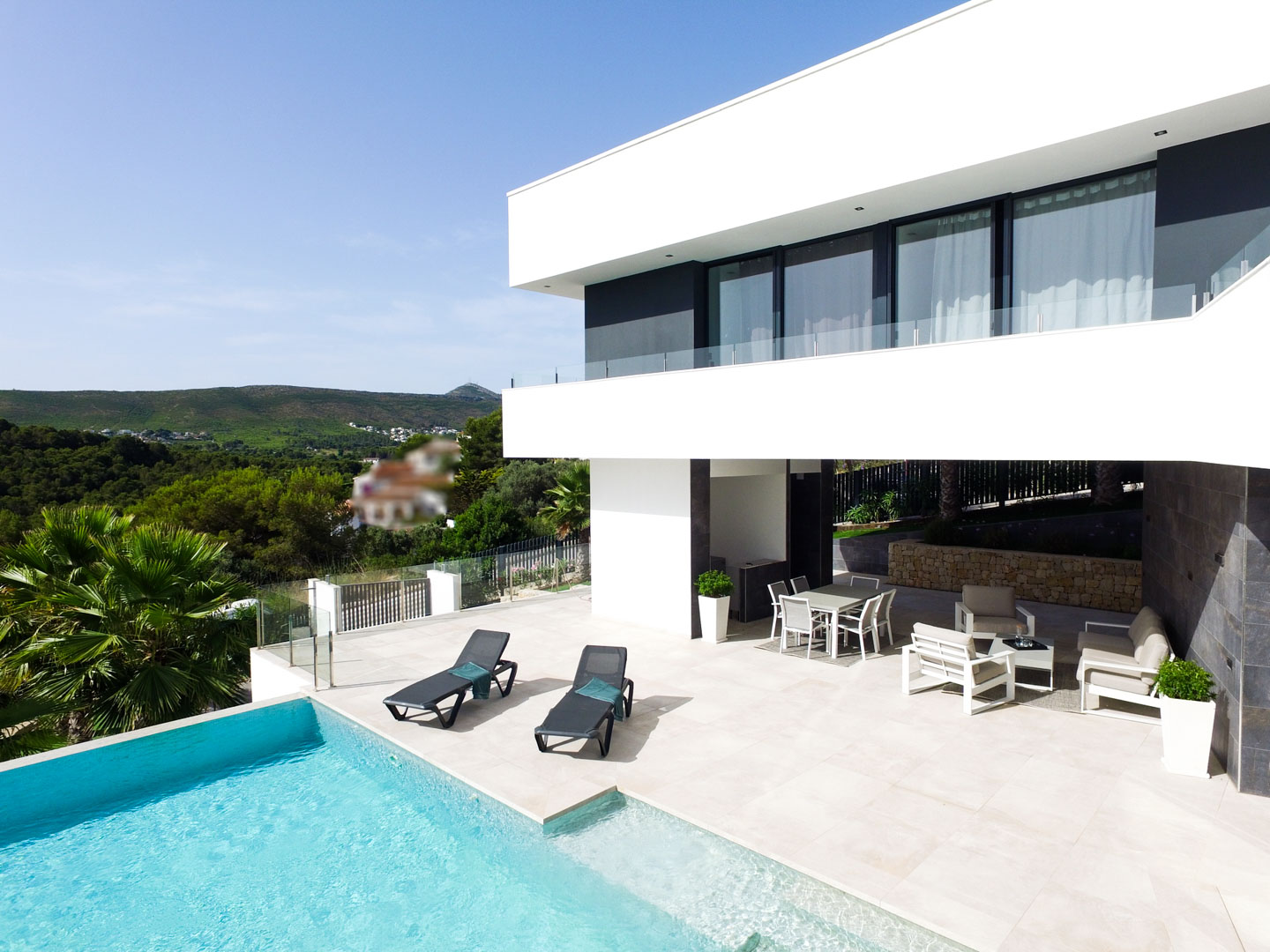 Main terrace featuring an infinity pool, sun loungers, and a covered lounge area with modern furniture set against a backdrop of green hills.