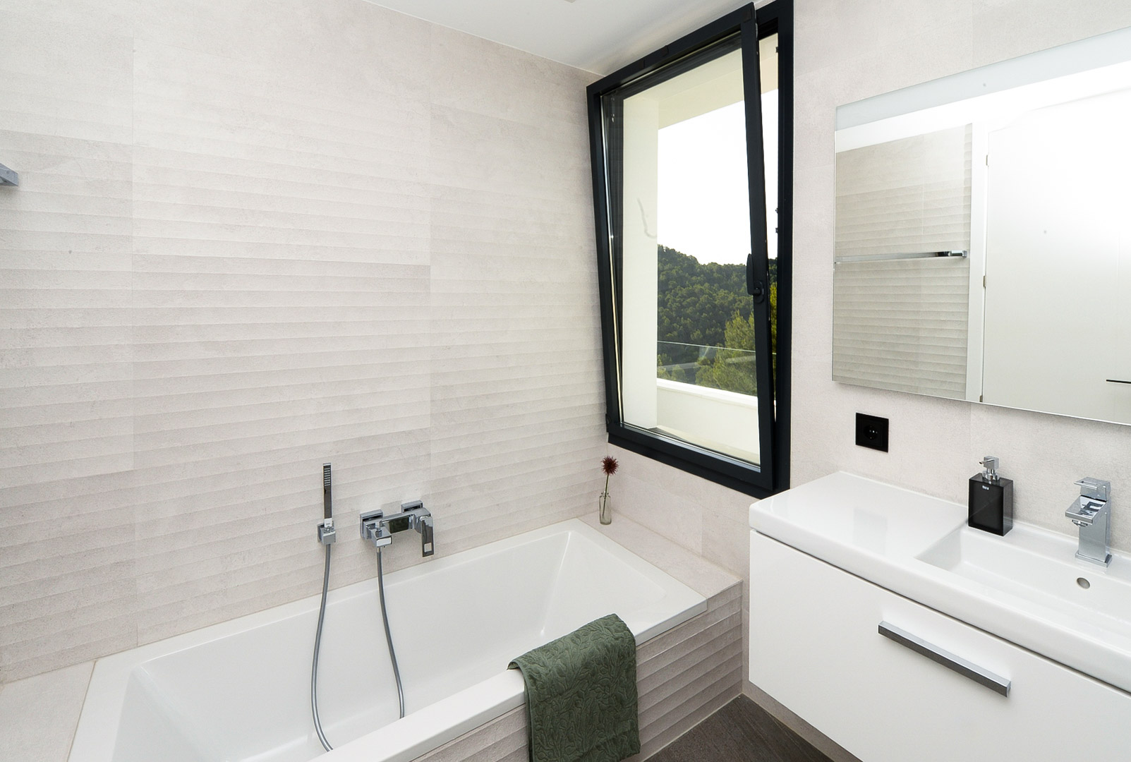 Modern bathroom featuring a built-in bathtub, textured wall tiles, and a window offering views of the surrounding Mediterranean greenery.