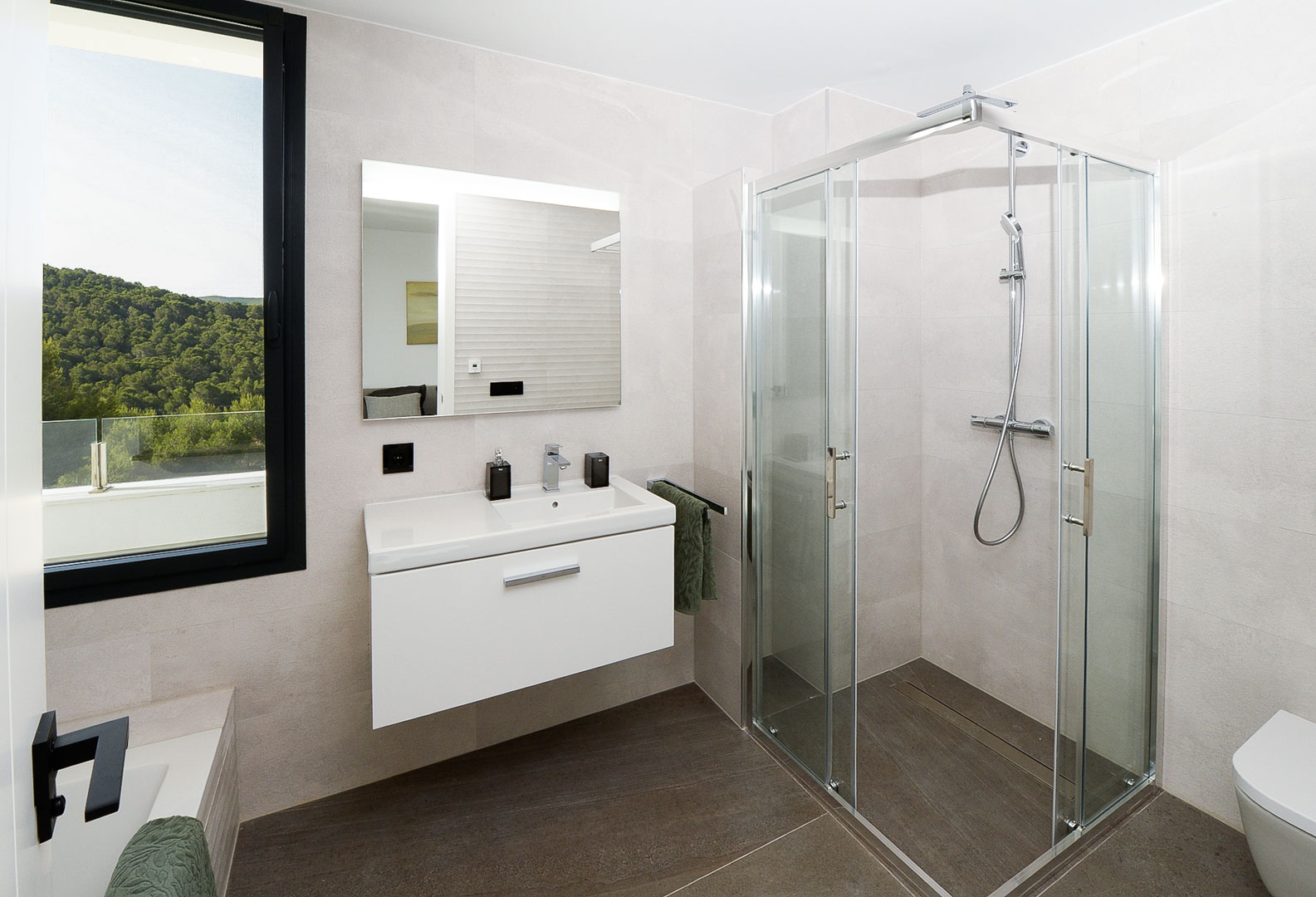 Modern bathroom featuring a glass shower cabin, floating white vanity, and a window overlooking green hills. Neutral wall tiles complement the dark flooring.