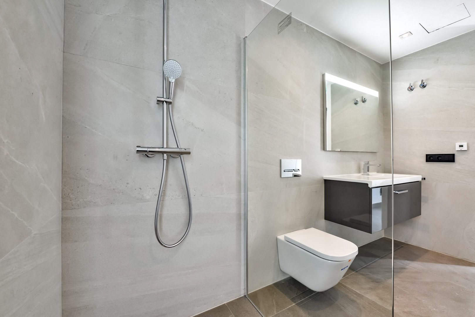 Modern bathroom featuring a walk-in shower with glass partition, wall-mounted toilet, and floating vanity. Large-format grey stone tiles and a backlit mirror complete the design.