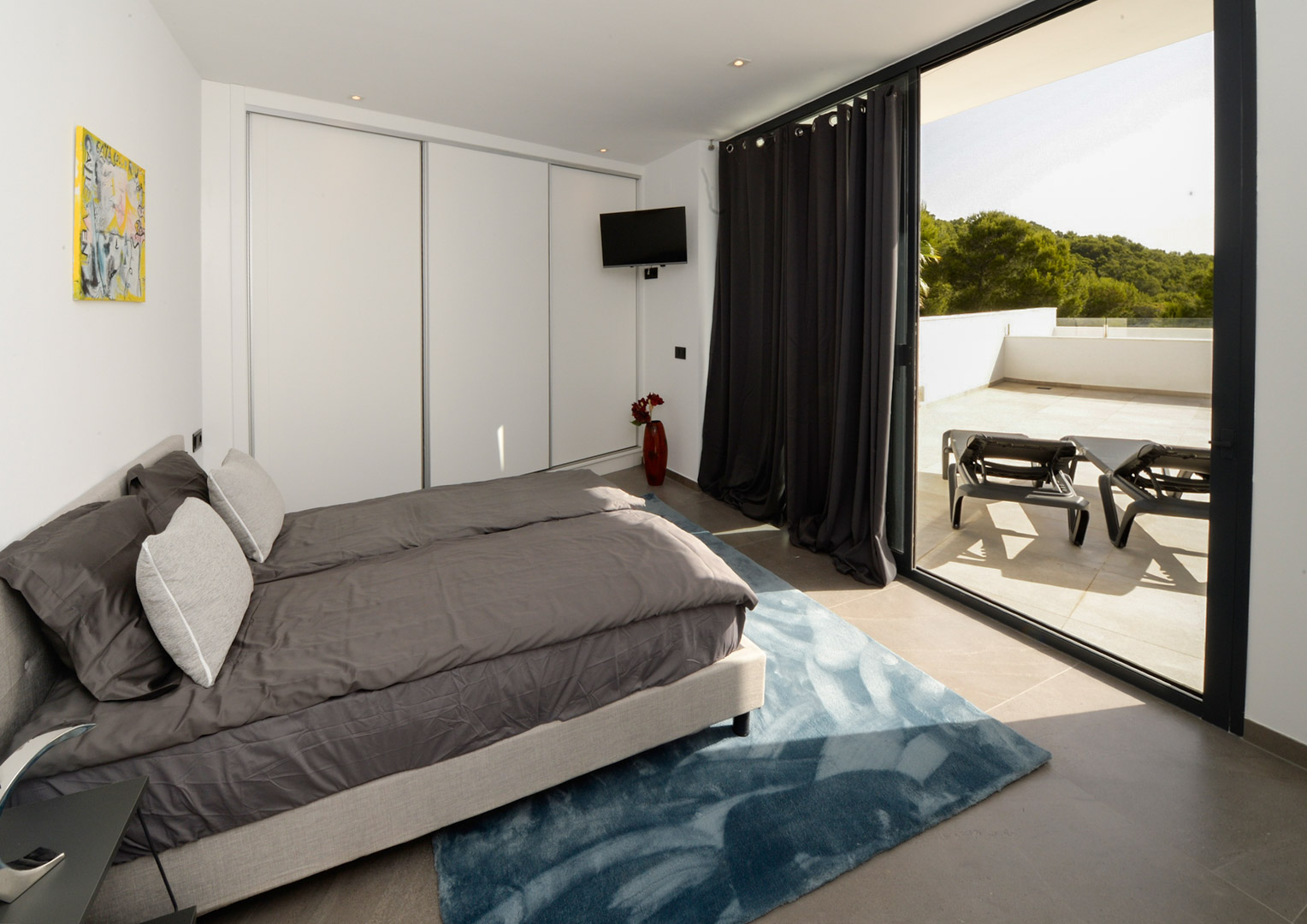 Modern bedroom featuring white built-in wardrobes and direct access to a spacious terrace with sun loungers and views of the surrounding greenery.
