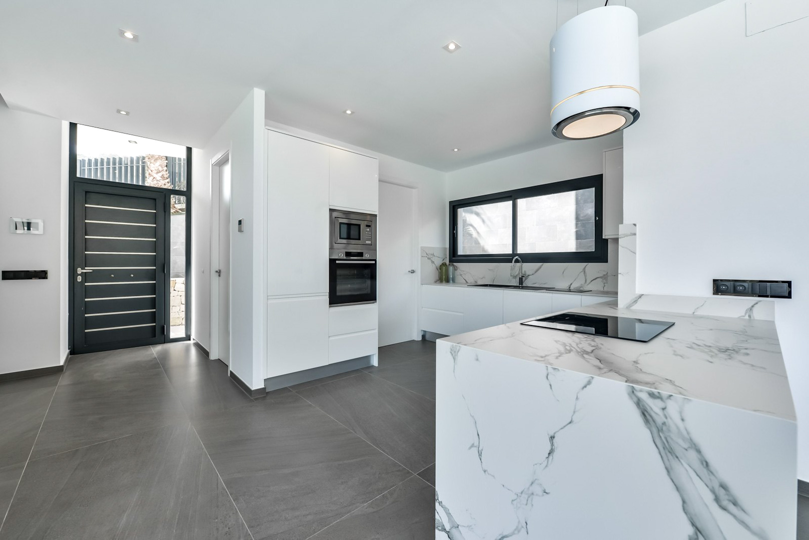 Modern designer kitchen featuring a marble-effect waterfall island, integrated appliances, and a cylindrical hood next to the black minimalist entrance door.