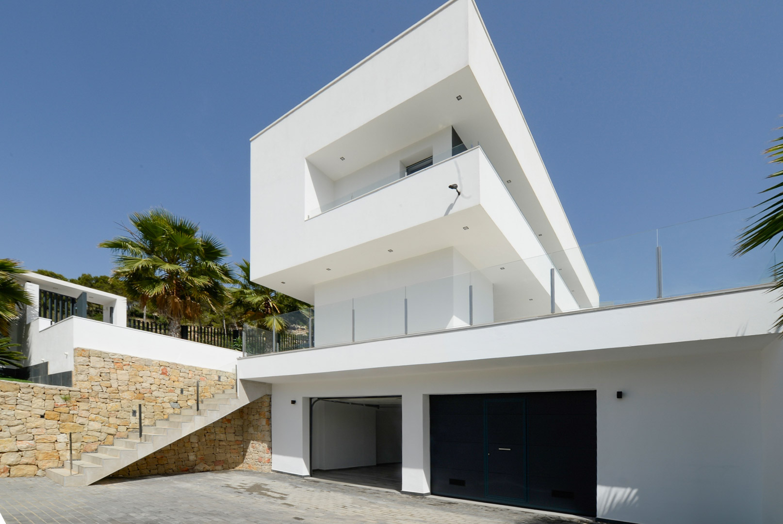 Modern exterior featuring white cubic volumes, a double garage, natural stone retaining walls, and outdoor stairs with glass balustrades.