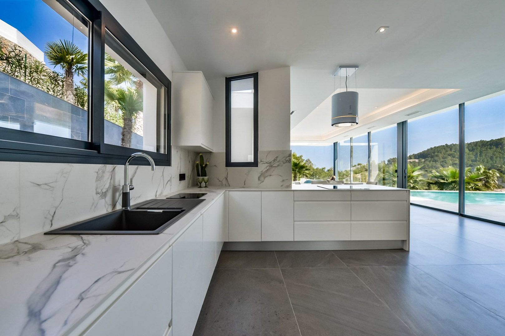 Modern kitchen featuring marble-effect countertops, a black sink, and a cylindrical extractor. Large glass walls offer panoramic views of the infinity pool and Mediterranean landscape.