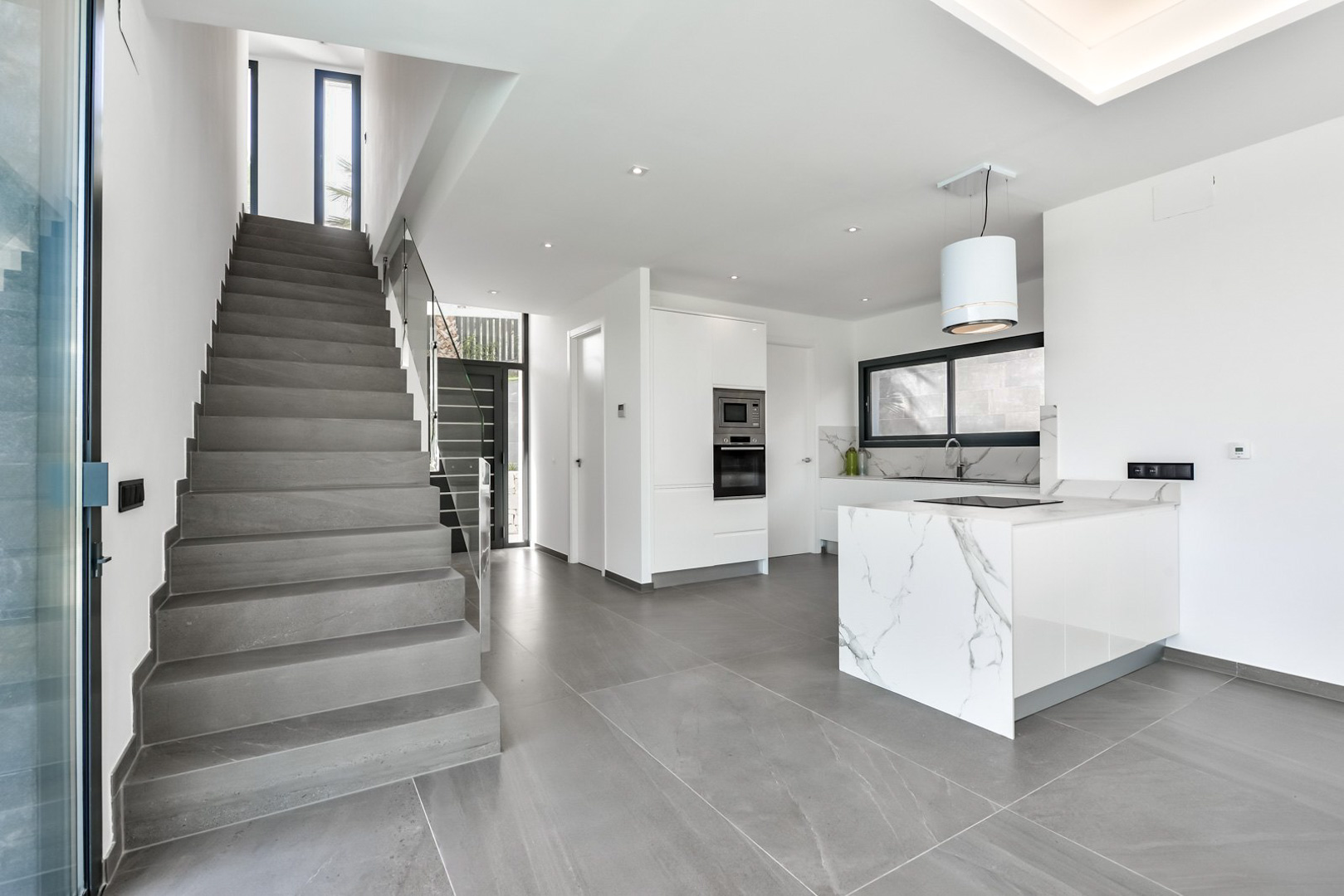 Modern open-plan kitchen featuring a marble waterfall island and integrated appliances, adjacent to a grey stone staircase with glass balustrades.