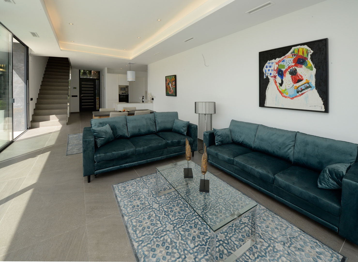 Open-plan living room featuring teal velvet sofas, a glass coffee table, and grey floor tiles. The space integrates the lounge with the dining area and modern staircase.