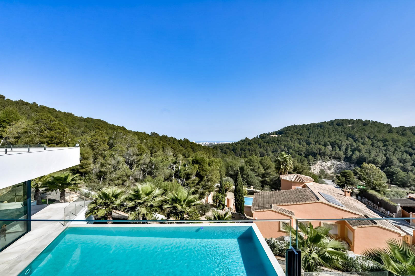 Panoramic view from the upper terrace featuring the infinity pool, glass balustrades, and expansive views over Mediterranean pine forests toward the sea.