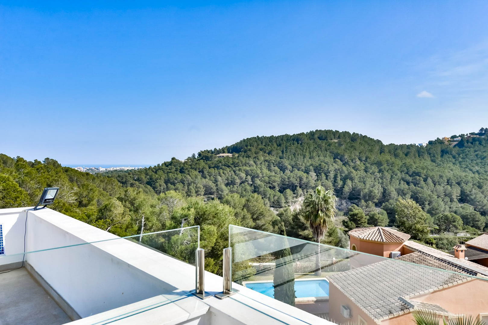 Upper terrace featuring a glass balustrade with stainless steel fittings, overlooking lush pine-covered hills and the distant Mediterranean coastline.