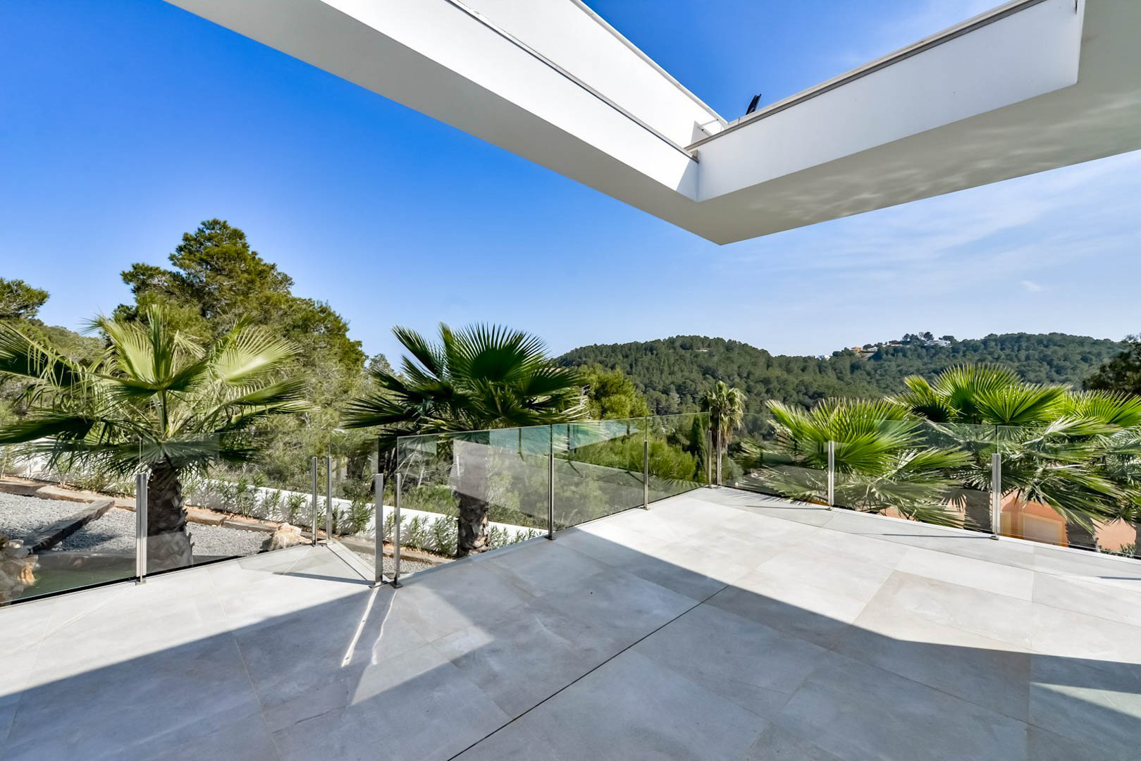Upper terrace featuring grey stone-effect tiling, glass balustrades, and panoramic views over the green hills and Mediterranean landscape.