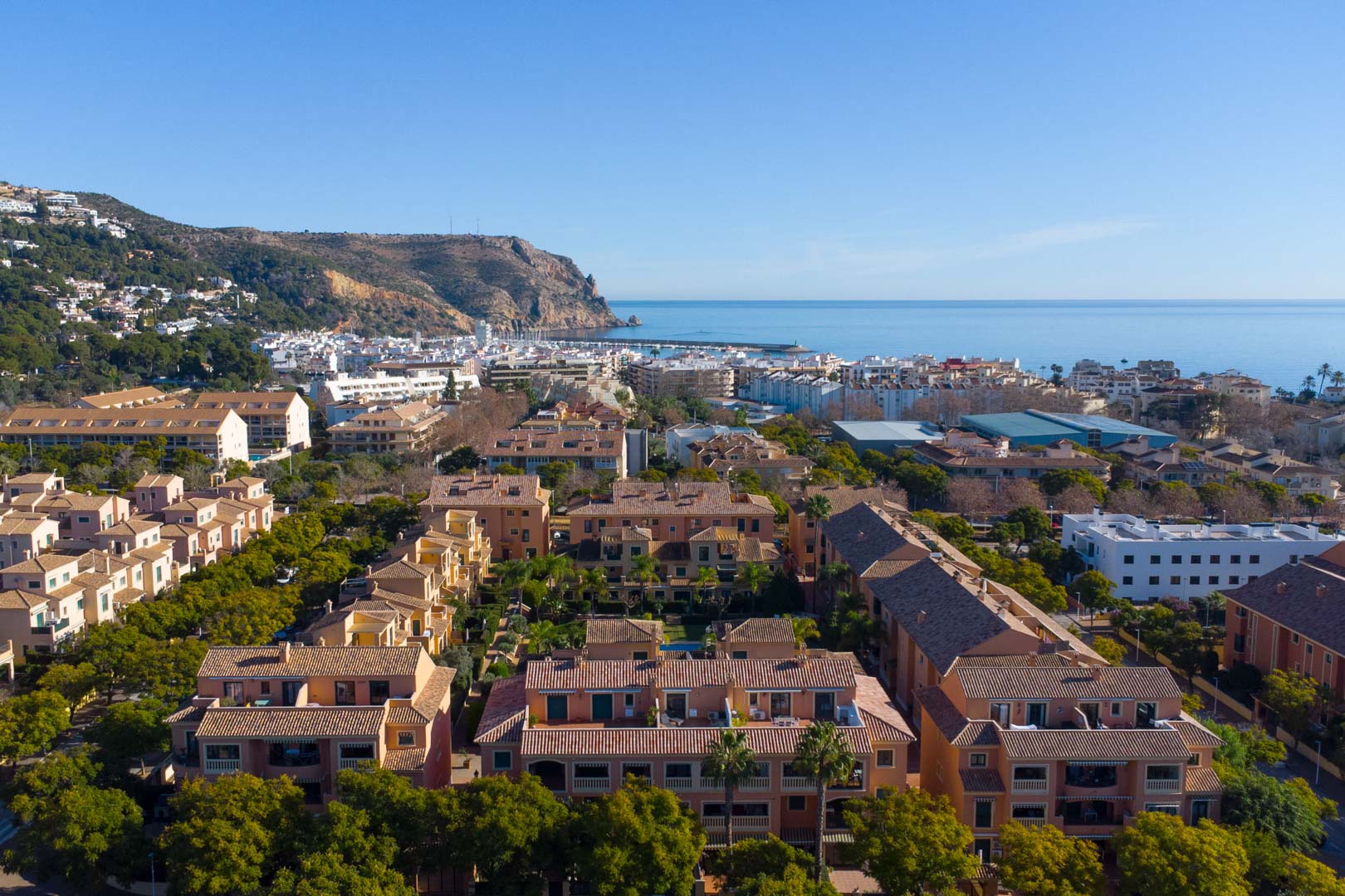Aerial view of the Floridamar complex in Jávea, featuring terracotta buildings and lush gardens with the Mediterranean Sea and port in the background.