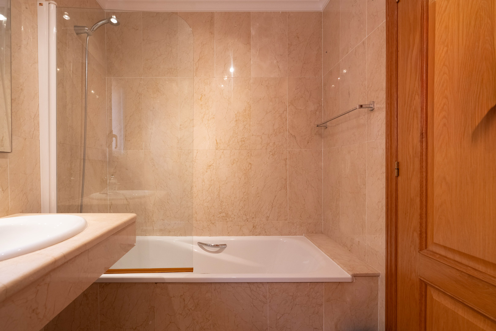 Bathroom featuring a bathtub with a glass partition and cream marble wall tiling. Includes a matching stone vanity and high-quality wooden door.