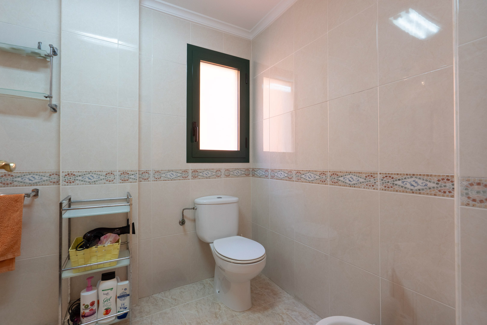 Bathroom featuring cream-toned wall tiles with a decorative mosaic border, a white toilet, and a frosted window providing natural light and ventilation.