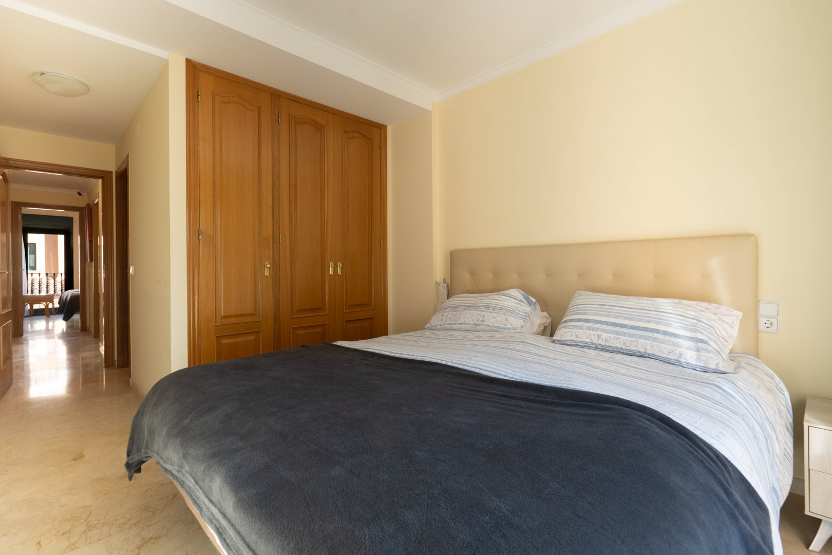 Bedroom featuring built-in wooden wardrobes, polished marble flooring, and a neutral tufted headboard, with a hallway leading to the living areas.