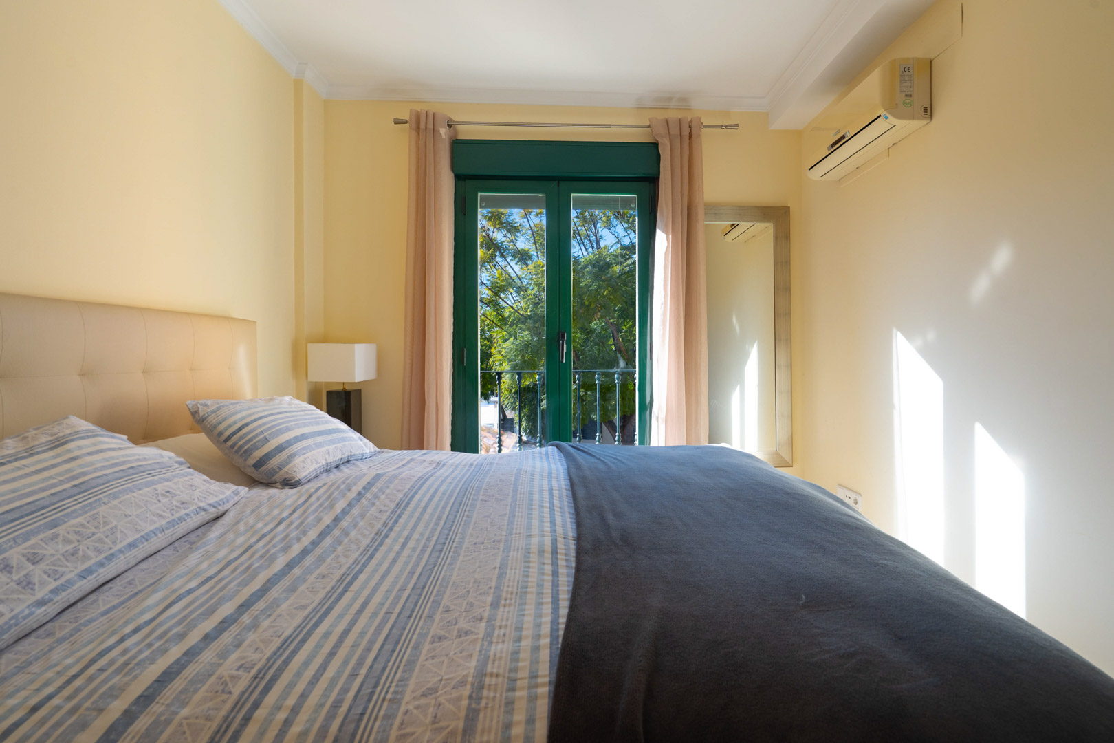 Bedroom with a double bed, padded headboard, and split air conditioning. Green balcony doors provide natural light and views of the greenery.