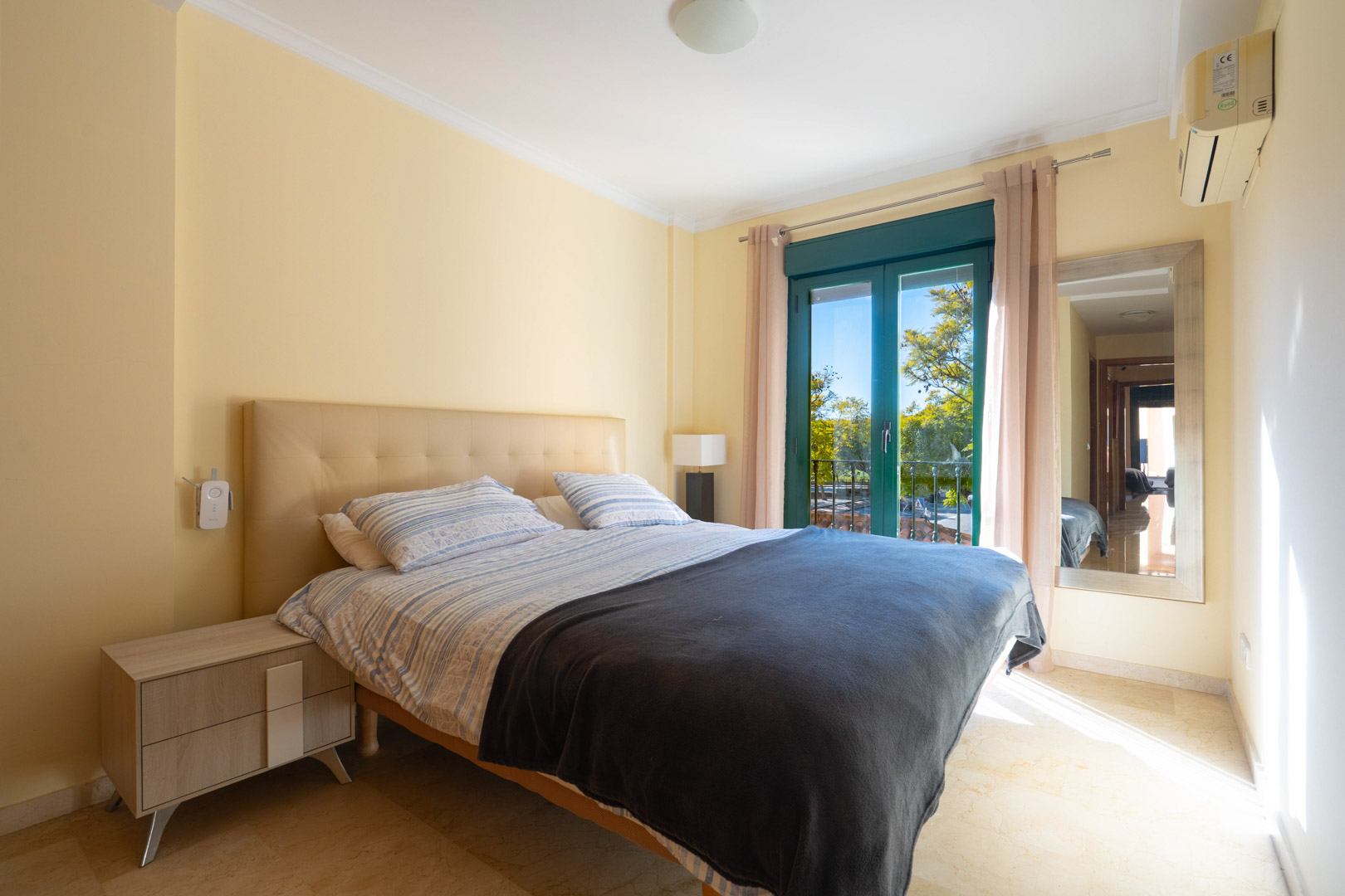 Bright bedroom featuring marble floors, a tufted headboard, air conditioning, and a balcony door offering views of the lush Mediterranean surroundings.