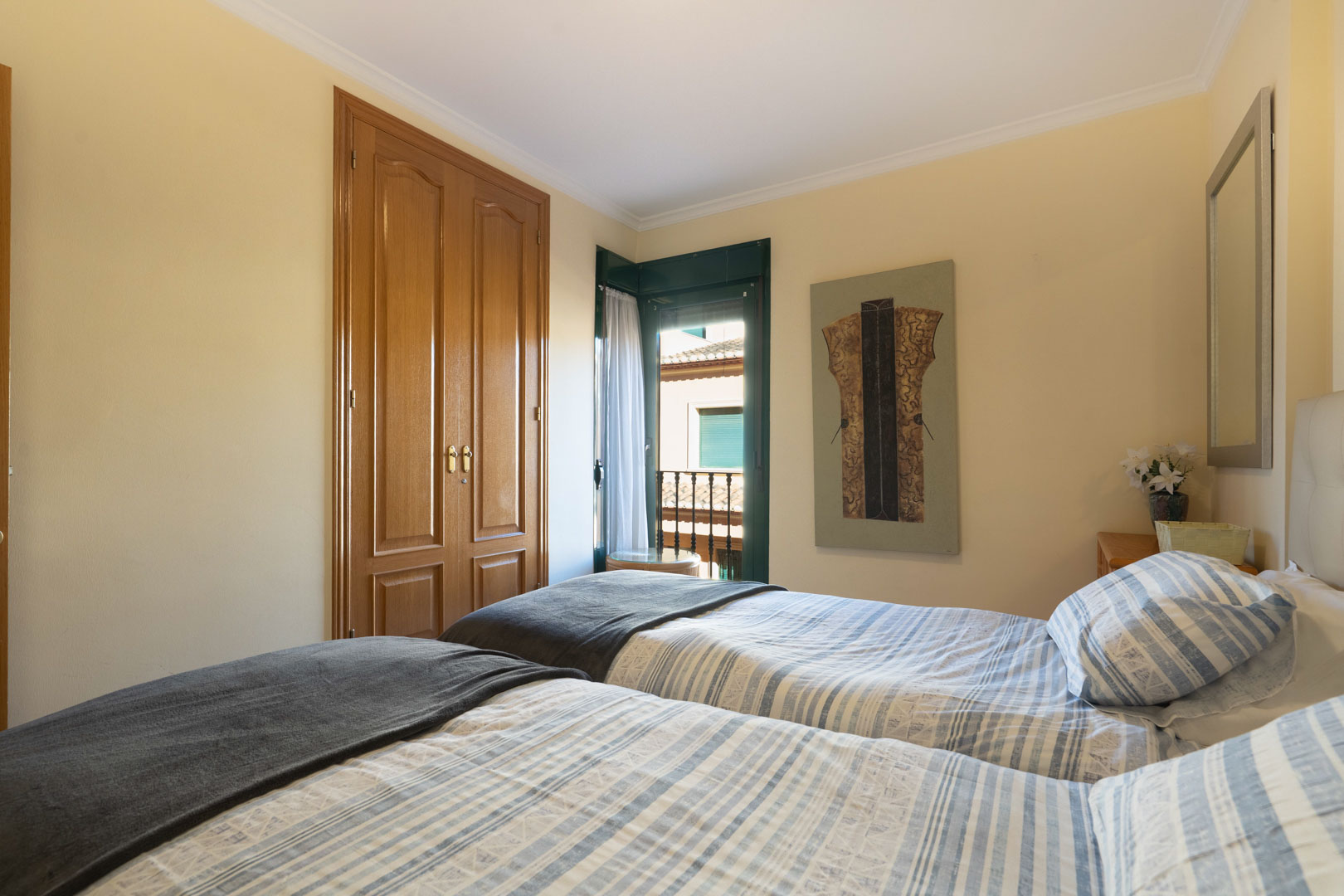 Bright bedroom featuring twin beds, built-in wooden wardrobes, and direct access to a balcony with traditional green shutters.