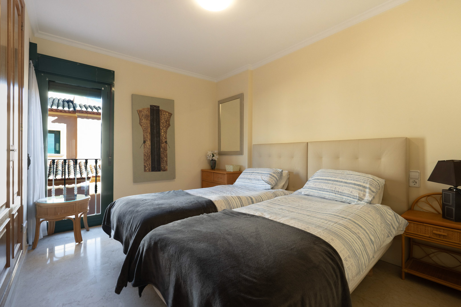 Bright bedroom featuring twin beds with a padded headboard, marble flooring, and built-in wardrobes. A glass door provides direct access to a private balcony.