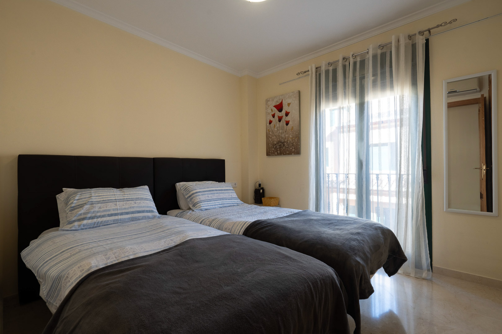 Bright bedroom featuring twin beds with dark headboards, polished marble flooring, and natural light from the balcony access.