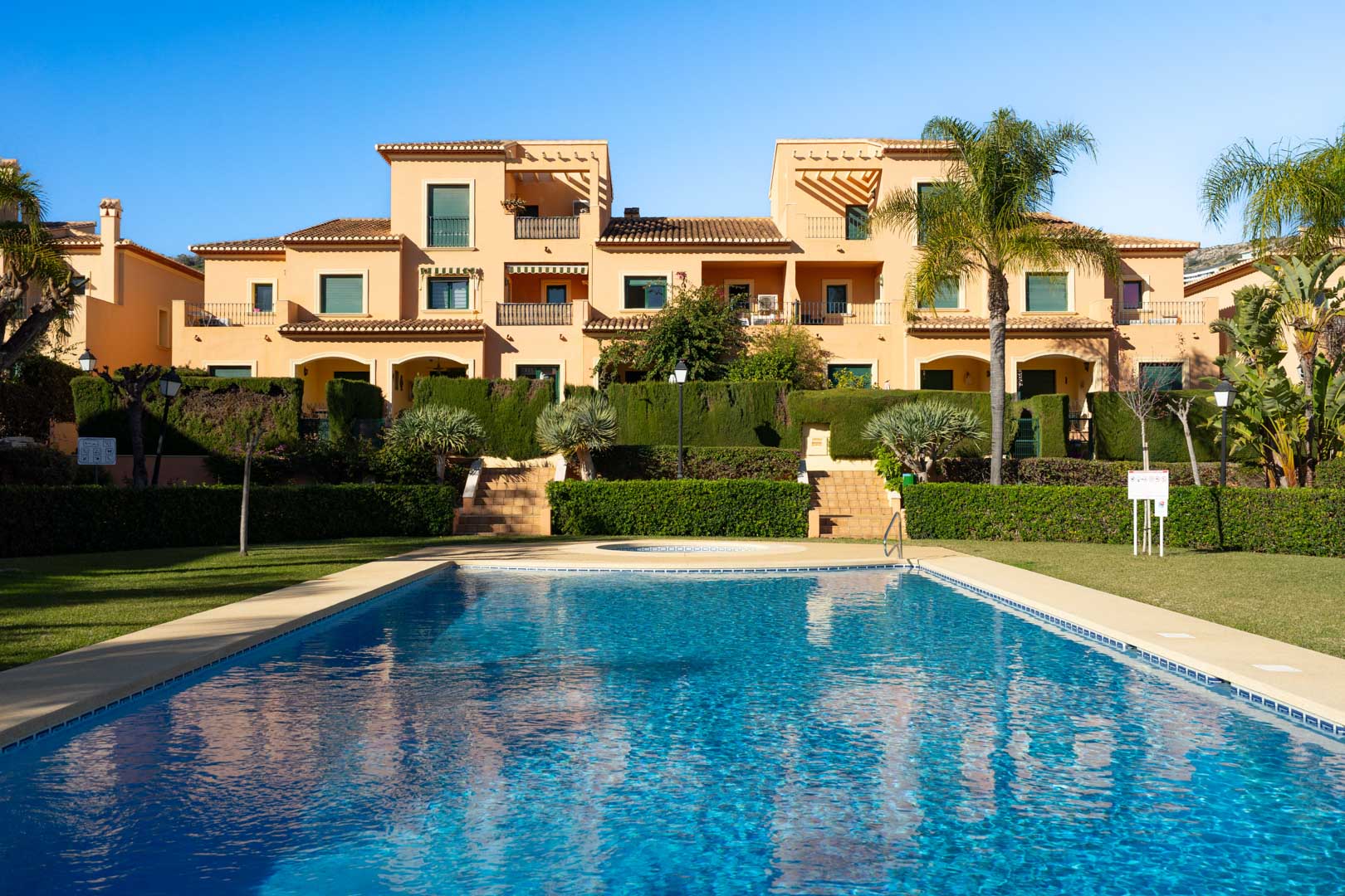 Communal swimming pool surrounded by manicured Mediterranean gardens and lawns, featuring the traditional architecture of the Floridamar complex in the background.
