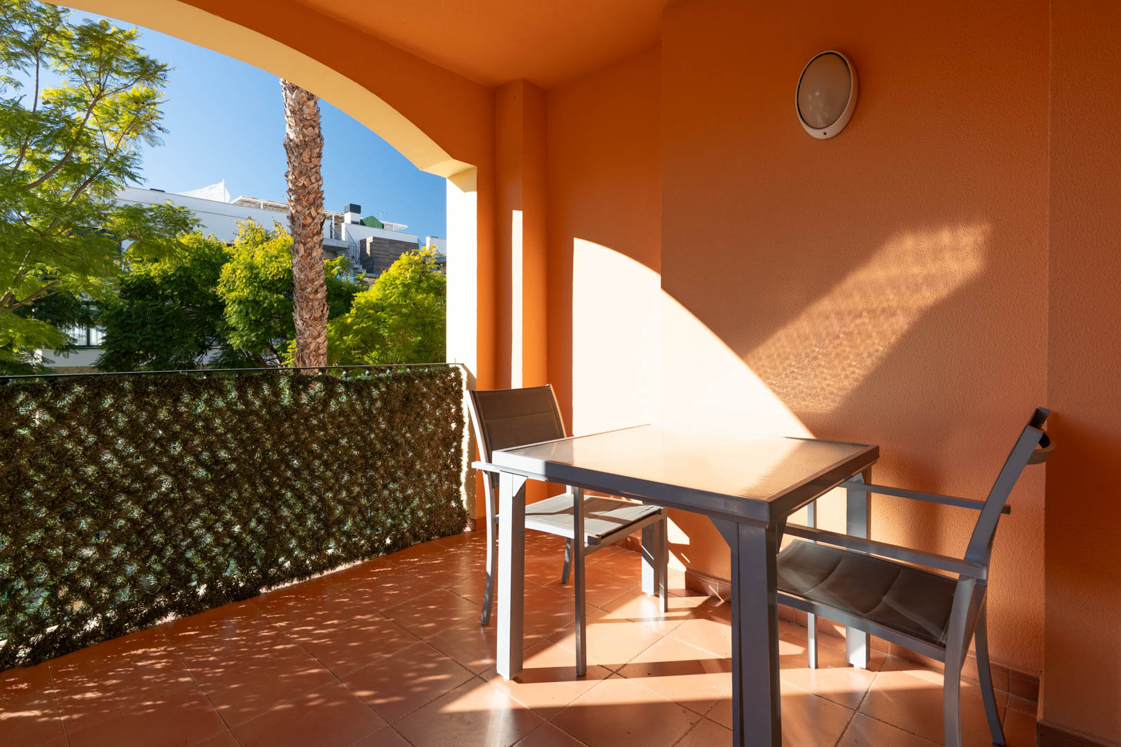 Covered terrace featuring terracotta tiles, a glass-top dining set, and views of the lush Mediterranean gardens and palm trees.