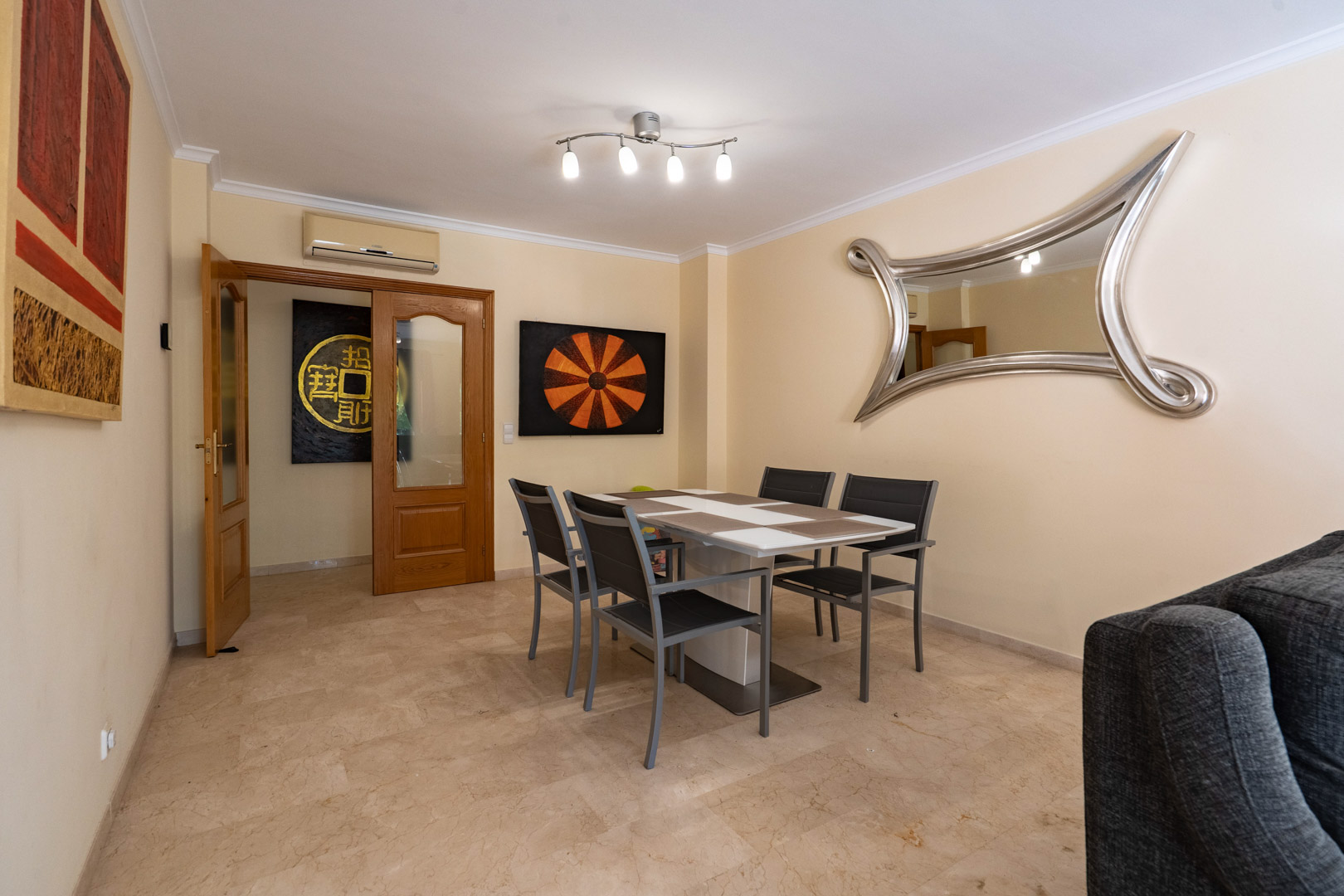 Dining area featuring marble floors, a modern four-seater table, and a large statement mirror, with wooden double doors leading to the hallway.