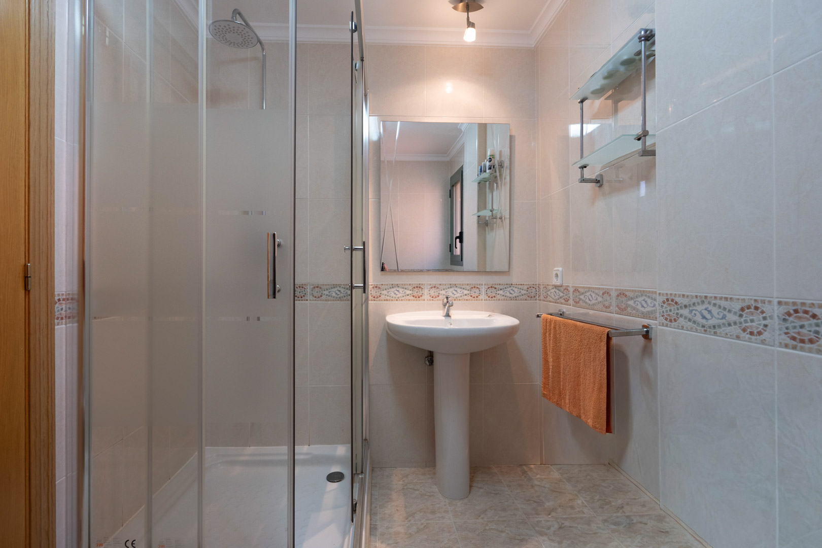 Functional bathroom featuring a glass-enclosed walk-in shower, pedestal sink, and tiled walls with a classic decorative mosaic border.