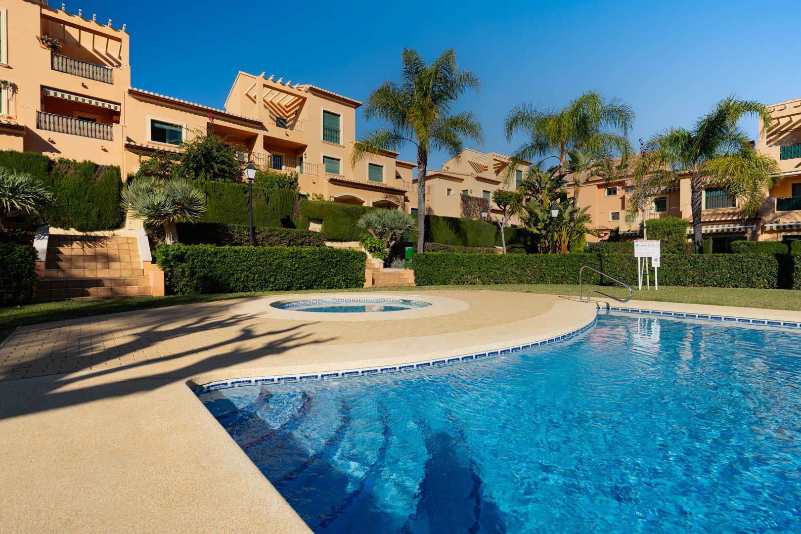 Large communal outdoor swimming pool featuring a curved design and circular plunge pool, set within landscaped gardens with mature palms and terracotta buildings.