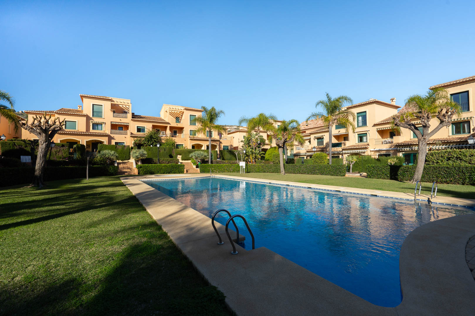 Large communal outdoor swimming pool set within manicured lawn gardens and palm trees, featuring Mediterranean-style architecture in the background.