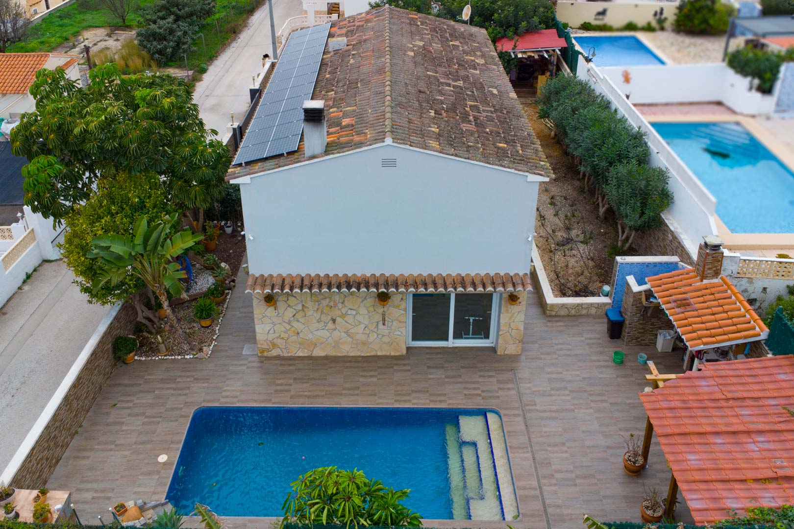 Aerial view of the villa featuring roof-mounted solar panels, a private swimming pool, and a paved terrace with a covered outdoor kitchen.