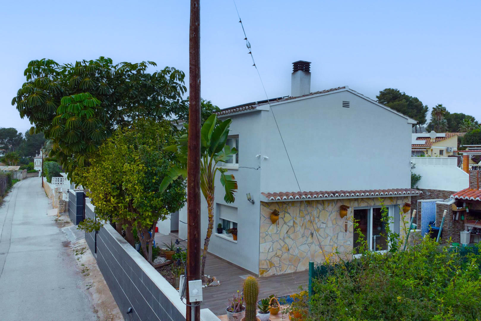 Exterior view of the villa showing the white facade with natural stone cladding, Mediterranean plants, and rooftop solar panels.
