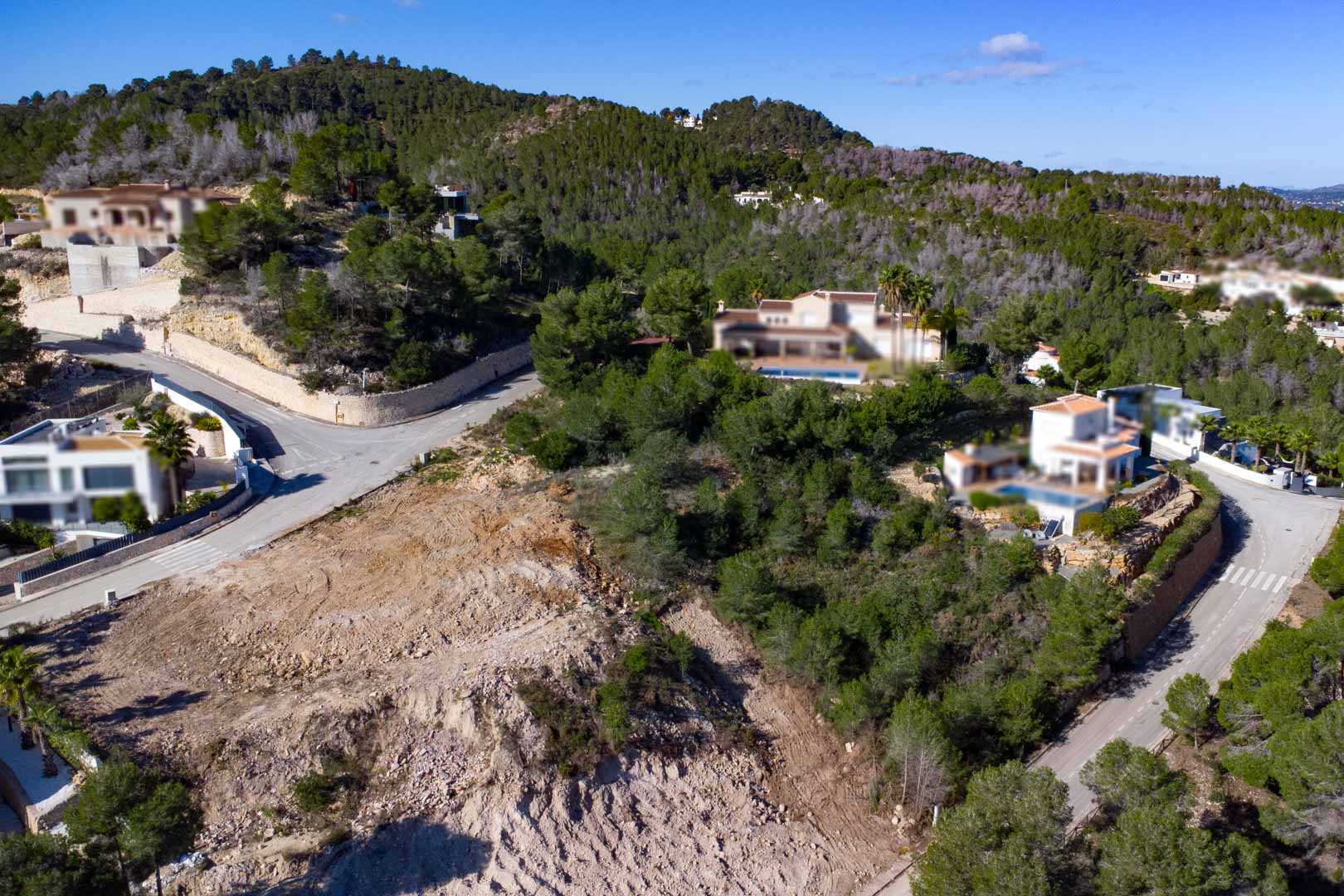 Elevated corner building plot in Tosalet 5, surrounded by pine forests and modern villas, featuring paved road access and a sloping terrain ready for construction.