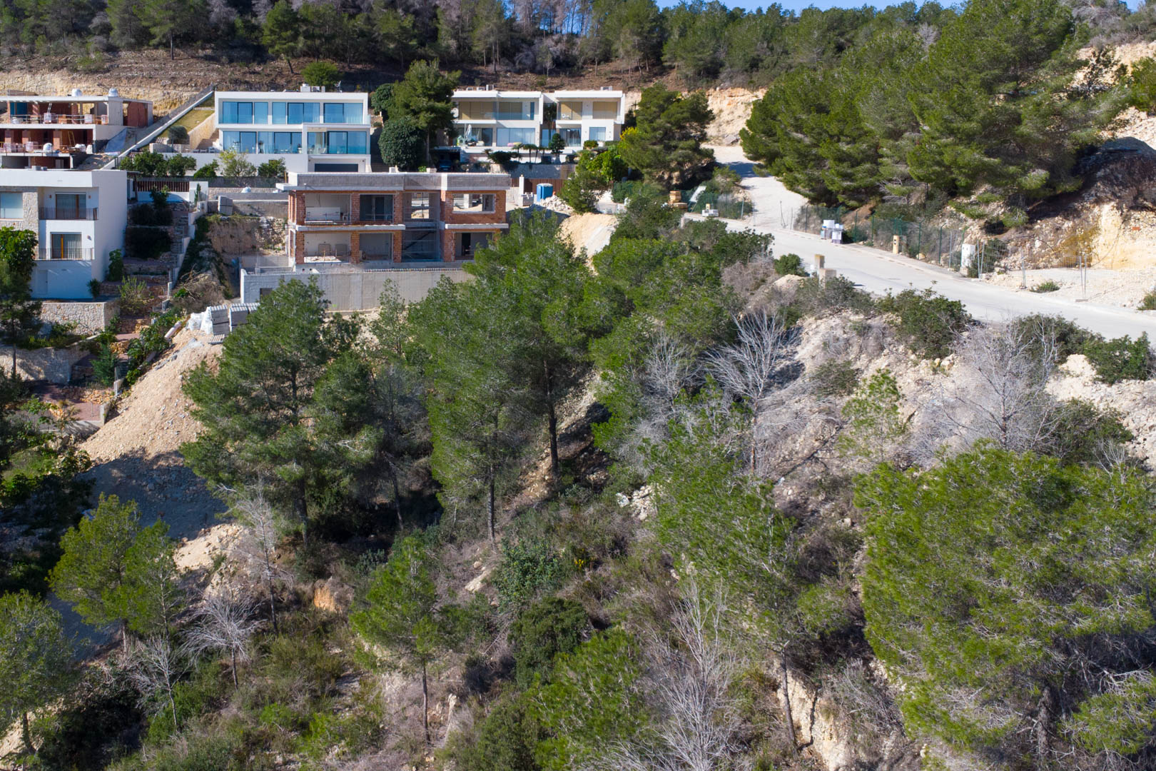 Elevated building plot in Tosalet 5, featuring Mediterranean pine trees and modern villas. The image shows the paved access road and a property under construction.
