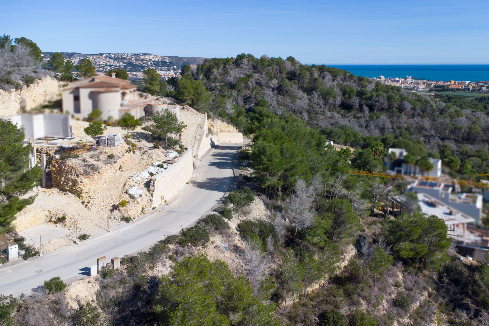 Elevated view of the paved access road in Tosalet 5, featuring stone retaining walls, Mediterranean pine forests, and distant views of the sea and coastline.