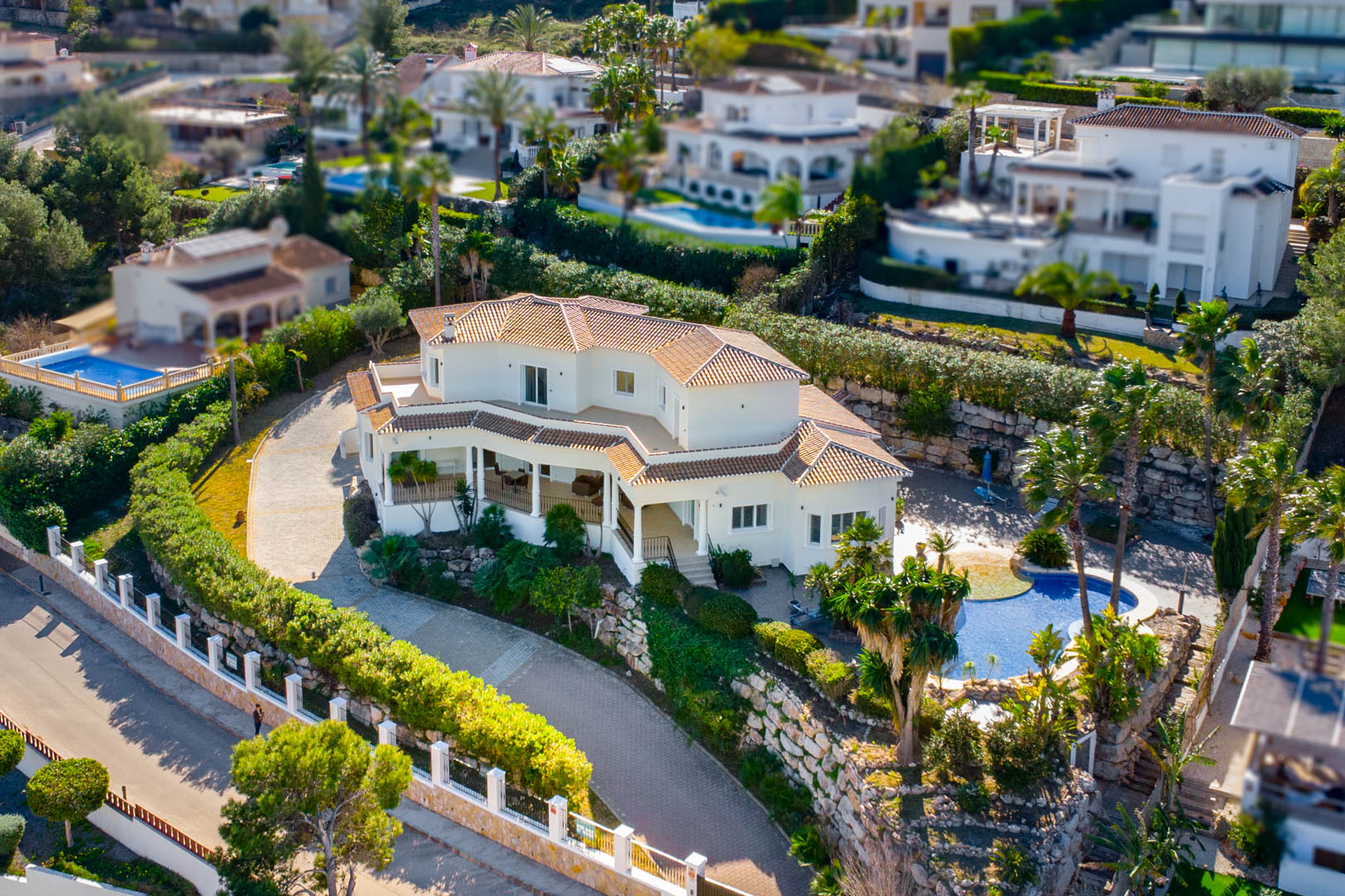 Aerial view of the villa featuring white facades, terracotta roofing, a curved stone driveway, and a private pool area surrounded by mature Mediterranean vegetation.
