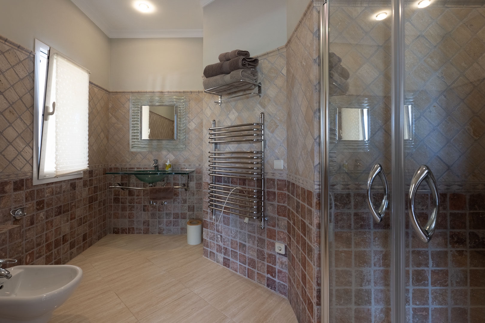 Bathroom featuring earthy-toned wall tiles, a glass vessel sink, chrome towel radiator, and a glass-enclosed shower cabin with recessed lighting.