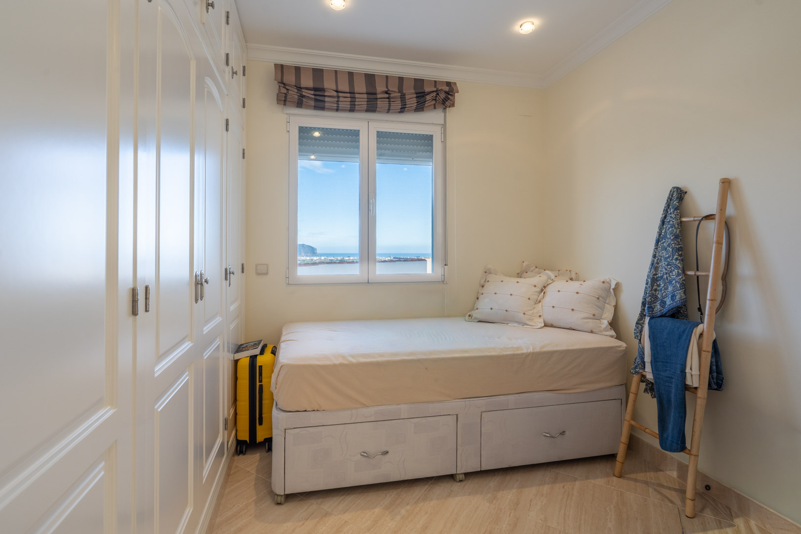 Bedroom featuring white built-in wardrobes, a bed with integrated storage drawers, and a window offering views of the Mediterranean coast.