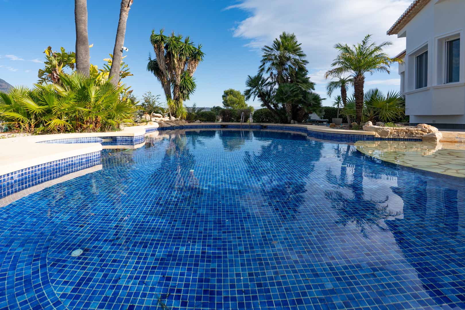 Blue mosaic swimming pool with an irregular design, surrounded by a tropical garden with palm trees and lush Mediterranean vegetation.