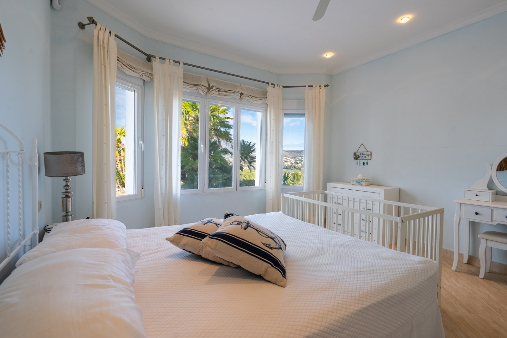 Bright bedroom featuring a bay window with views of the Montgó, white furniture including a crib and vanity, and nautical-themed decor.