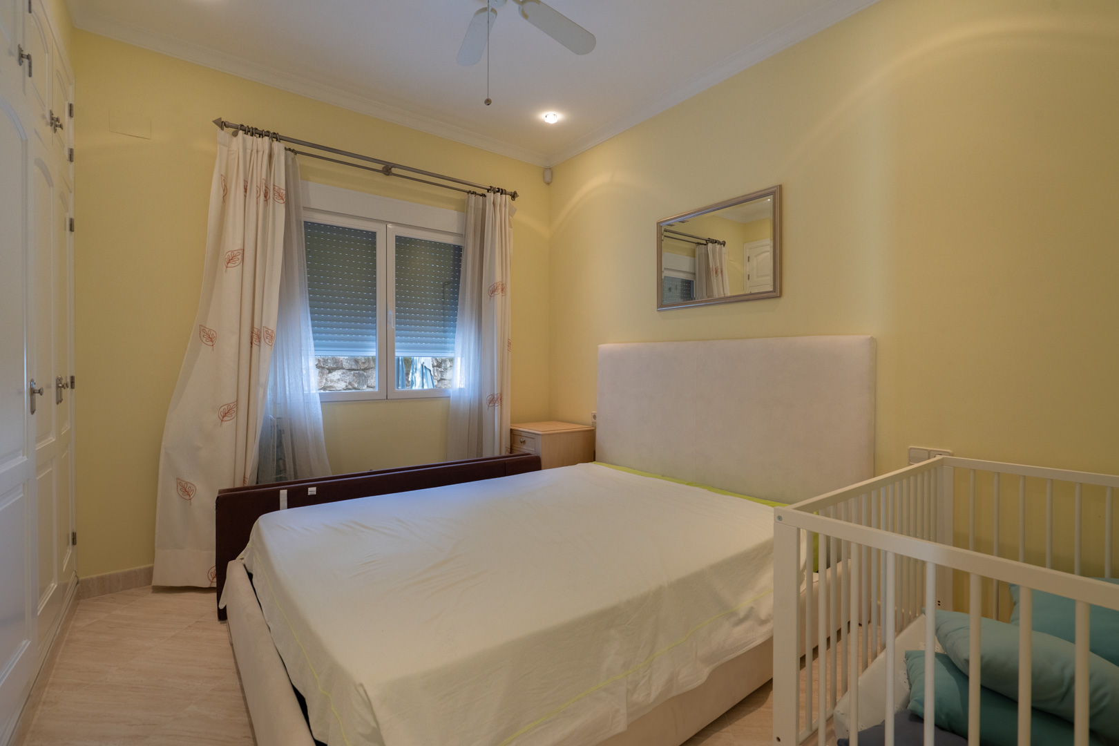 Bright bedroom featuring cream-colored walls, white built-in wardrobes, a double bed with an upholstered headboard, and a crib. Equipped with a ceiling fan and recessed lighting.